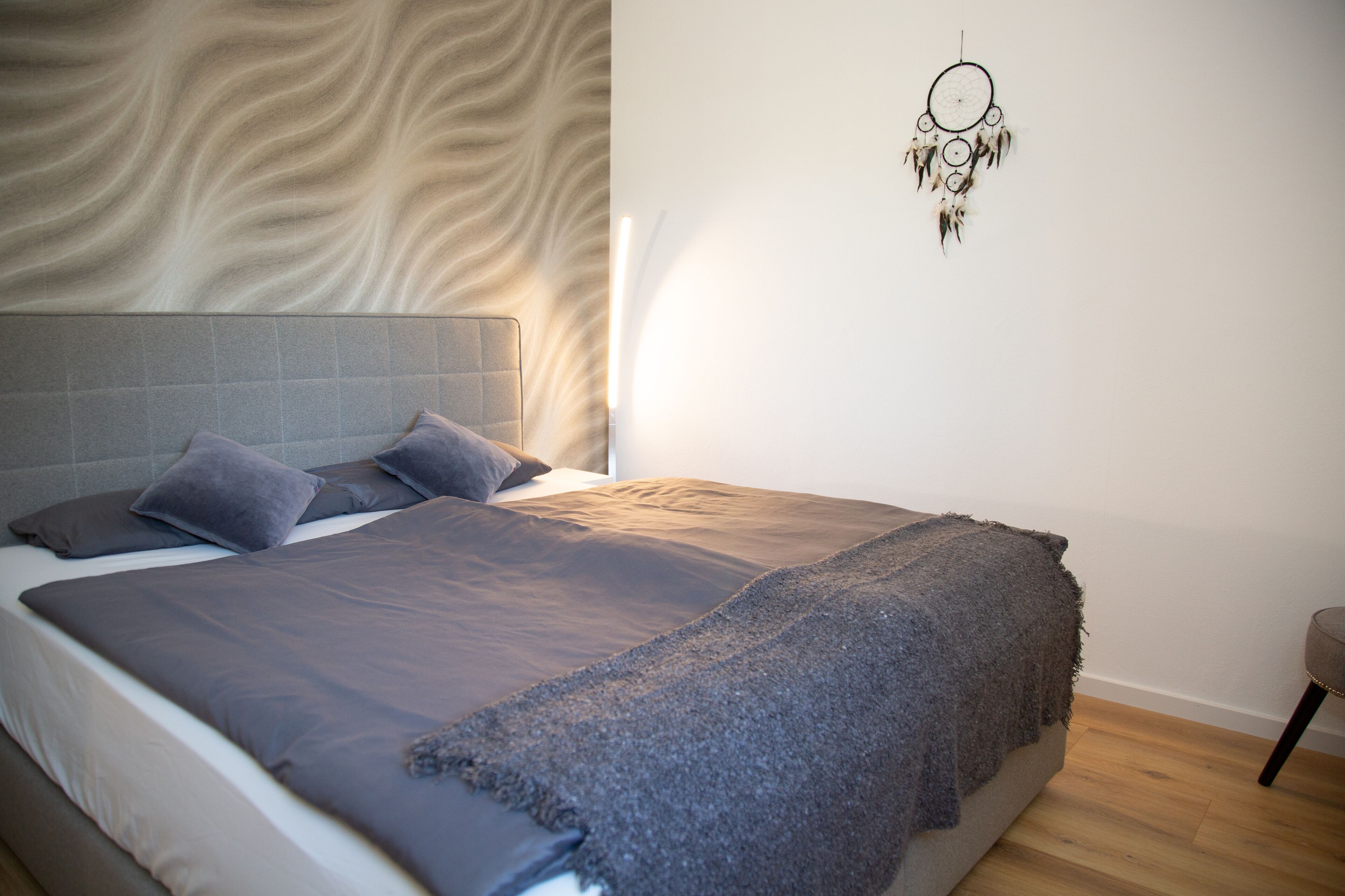 Un lit bien fait, avec une couverture et des oreillers gris, une tête de lit ornée d’un motif ondulé et un attrape-rêves accroché au mur.