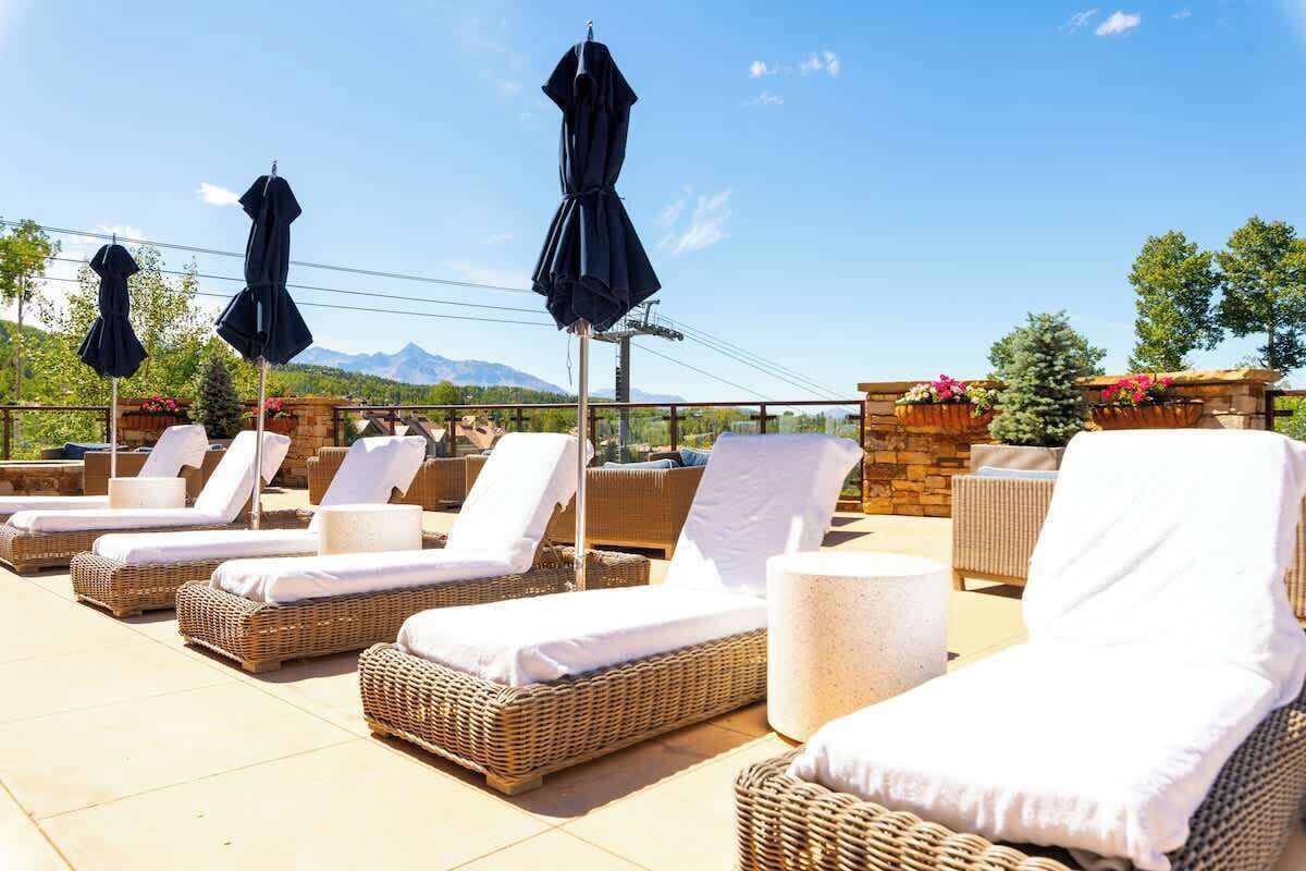 A rooftop lounge area with wicker lounge chairs and umbrellas, a stone wall with flower boxes, and a view of mountains.