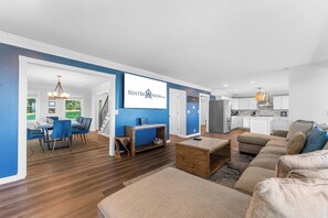 A modern living room with a dining area, kitchen, and a large screen displaying a logo. - Ultimate Lakeview w/ Private Outdoor Pool, Hot Tub, Yard, Karaoke, Billiards, Arcade & Free Tickets (Branson)