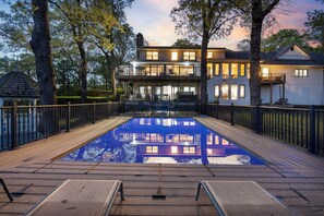A swimming pool with a wooden deck and metal railings, surrounded by trees and a two-story building with balconies.