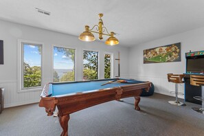 A pool table with a wooden frame and a blue felt surface, a chandelier with three lights, and a painting of a pool game on the wall. - Ultimate Lakeview w/ Private Outdoor Pool, Hot Tub, Yard, Karaoke, Billiards, Arcade & Free Tickets (Branson)
