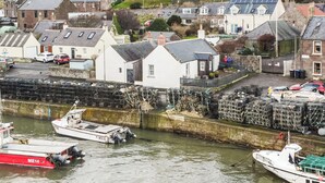 A harbor with boats docked, a white building with a grey roof, and a stone wall with stacked crates.