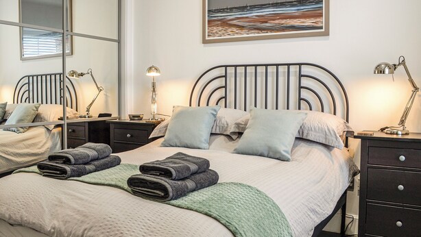A neatly made bed with pillows and folded towels, a bedside table with a lamp, a mirror, and a painting on the wall.