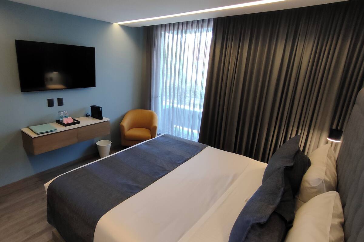 A modern hotel room with a large bed, a flat-screen TV mounted on the wall, a small desk with a chair, and a window with curtains.