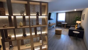 A modern living room with a sectional sofa, a round ottoman, a coffee table, and a TV mounted on the wall. There is a decorative shelf with various items and a window with curtains. - Alia Inn Boutique WTC (Mexico City)