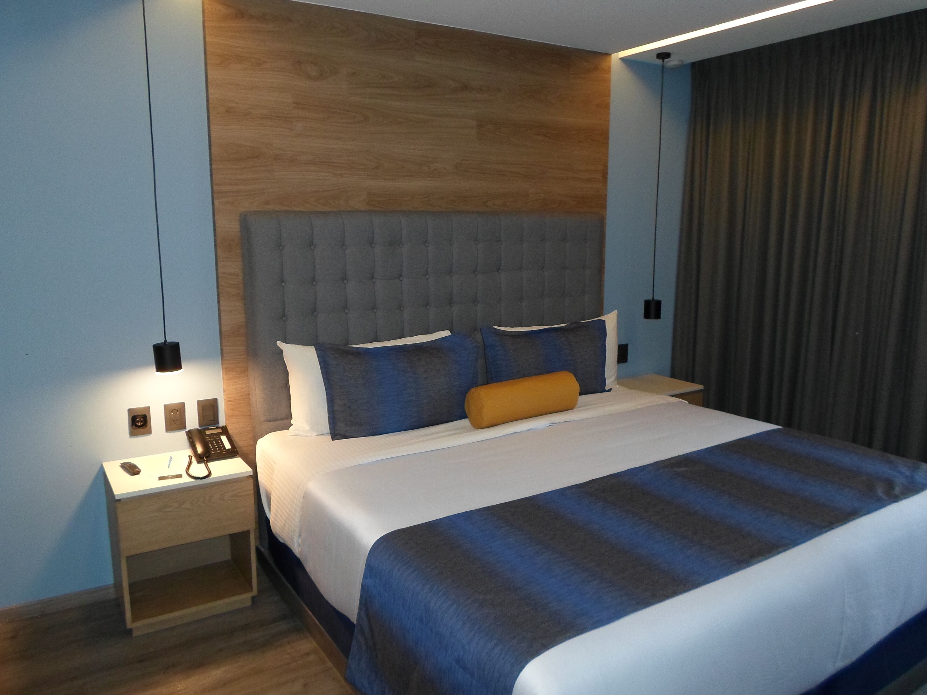 Ein modernes Hotelzimmer mit einem großen Bett, einem Holzkopfstück, einem Nachttisch mit Telefon und Lampe sowie einem Vorhang am Fenster.