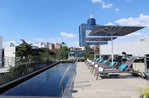 Un área de piscina en la azotea con sillones de descanso y sombrillas, una vista despejada del horizonte de la ciudad y un edificio moderno al fondo.