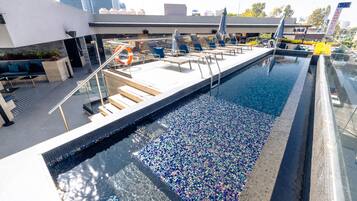 Una piscina en la azotea con piso de mosaicos, sillas de descanso y un flotador.