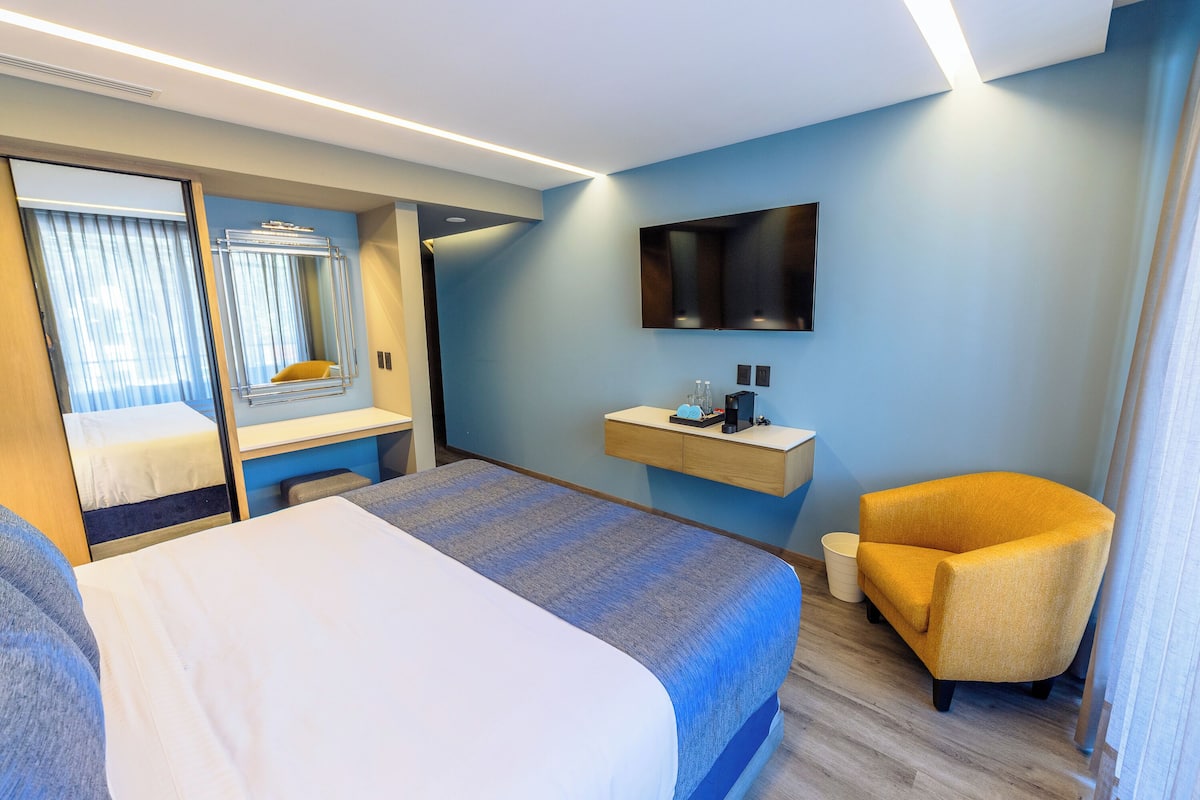 A hotel room with a bed, a chair, a desk, a mirror, and a mounted TV.