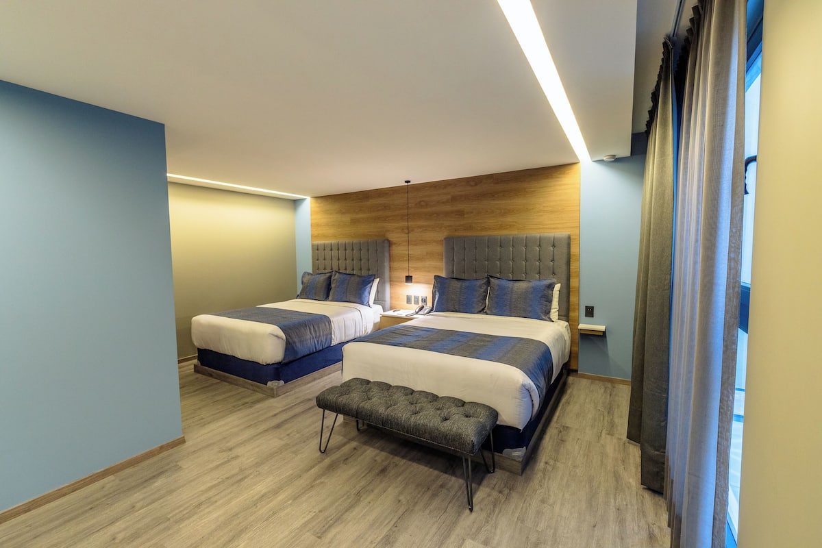 A hotel room with two beds, a bench, a wooden headboard, and a nightstand.