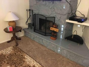 Cozy room features a fireplace, a small table with a lamp and a box, and a shelf with electronic devices.