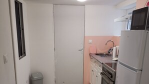 A compact kitchen with a microwave, oven, and sink. A trash bin is against the wall, and a door leads to another room.