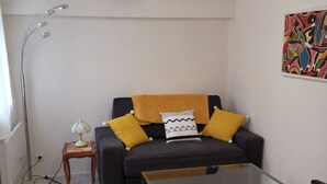 A living room with a black sofa, yellow cushions, a glass coffee table, a wooden side table with a lamp, and a colorful abstract painting on the wall.