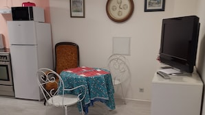 A small, well-lit room with a dining table, chairs, a clock, and a television on a stand.