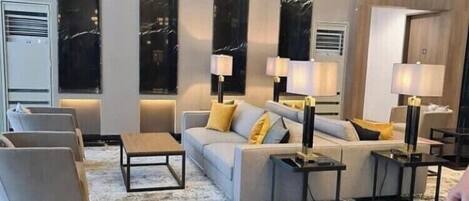 Una sala de estar moderna con un sofá, sillas y una mesa de centro. Hay lámparas de pared y un ventanal con vistas.