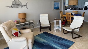 A living room with a beige sofa, two white armchairs, a small side table, a blue rug, and a kitchen in the background. - Expansive Ocean front views—direct beach access! (New Smyrna Beach)