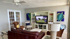 A living room with a brown leather sofa, a television, a ceiling fan, and decorative wall art. - Expansive Ocean front views—direct beach access! (New Smyrna Beach)