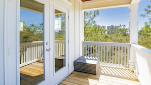 A covered porch with a wooden deck, white railings, and a view of trees and buildings in the background.
