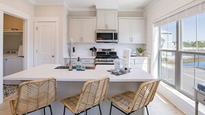 A modern kitchen with white cabinetry, a central island with bar stools, stainless steel appliances, and a large window offering a view of the outdoors.