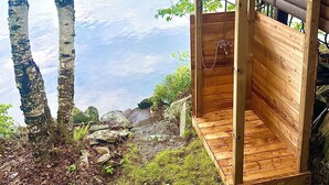 A wooden deck with a bench overlooking a calm lake, surrounded by birch trees and a wooden structure with a glass enclosure.