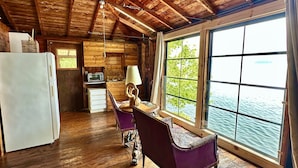 A rustic cabin interior with wooden floors, a dining area with a table and chairs, a kitchen with a refrigerator, and a large window overlooking a body of water.