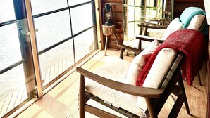 A wooden cabin with a large window overlooking a body of water, a wooden chair with cushions, and a small table.