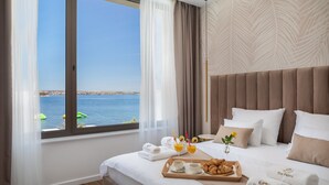 Habitación de hotel con un amplio ventanal con vista al mar, cama con ropa blanca, bandeja con croissants y jugo, y toallas con el logotipo del hotel.