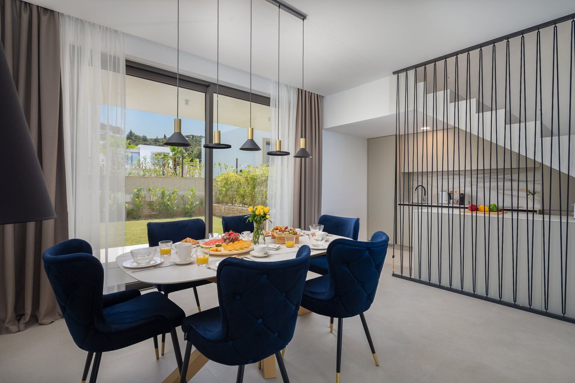 A modern dining area with a large table, blue chairs, and a view of a garden through large windows.