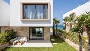 Una casa moderna de dos pisos con fachada de piedra, puerta de cristal y un patio con mobiliario exterior, y vista al mar.