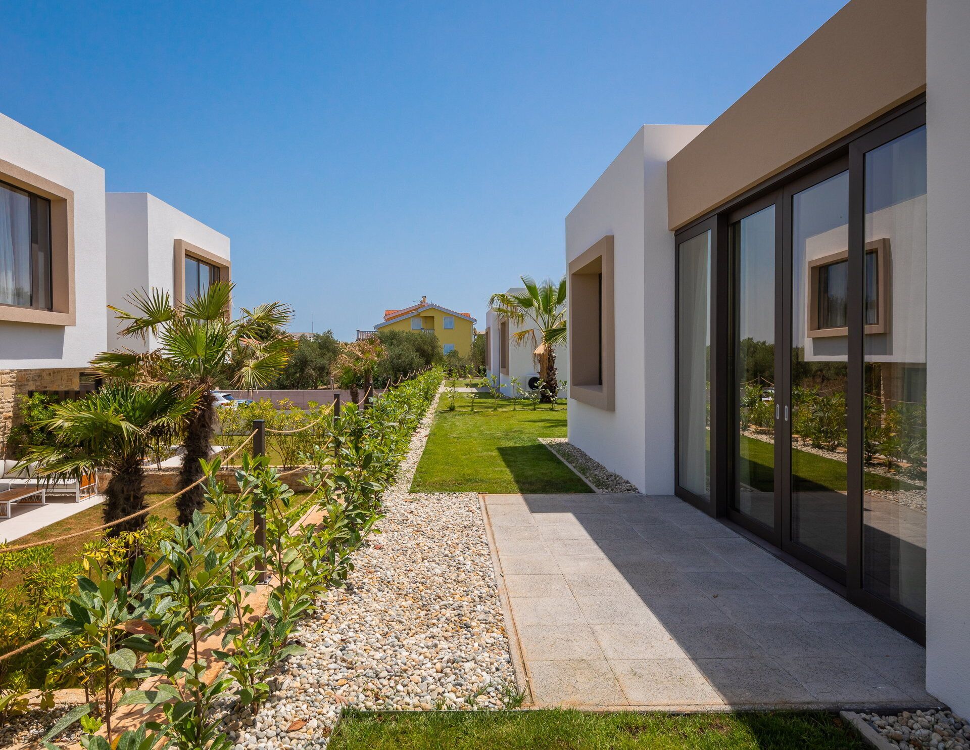 Una villa moderna con un sendero pavimentado, abundante vegetación y un cielo azul despejado.