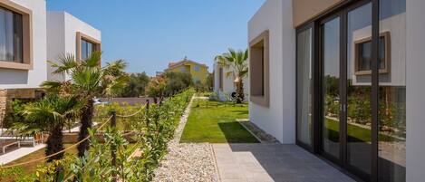 Una villa moderna con un sendero pavimentado, abundante vegetación y un cielo azul despejado.