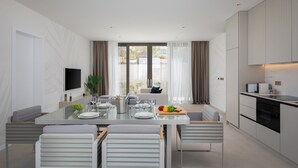 A modern kitchen with a dining area, featuring a large table with chairs, a TV mounted on the wall, and a view of the outdoors through the windows. - The Palms Resort - Villa Dream IV (Tkon)