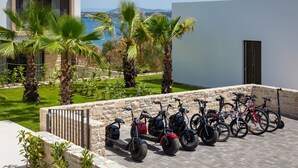 Een dakterras parkeergebied met elektrische scooters en fietsen, omgeven door palmbomen en uitzicht op de zee.