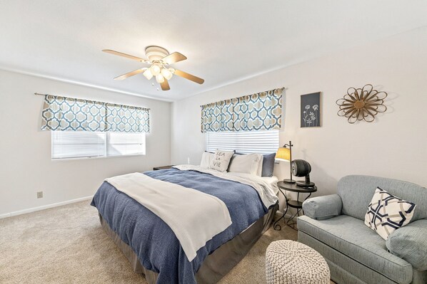 A bedroom with a bed, a gray sofa, a ceiling fan, patterned window valances, and a wall-mounted decor piece.
