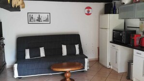 A living room with a sofa, a wooden coffee table, a hanging light fixture, a clock, and a kitchen area with appliances. - Town house in the heart of a typical Lot village (Saint-Sozy)