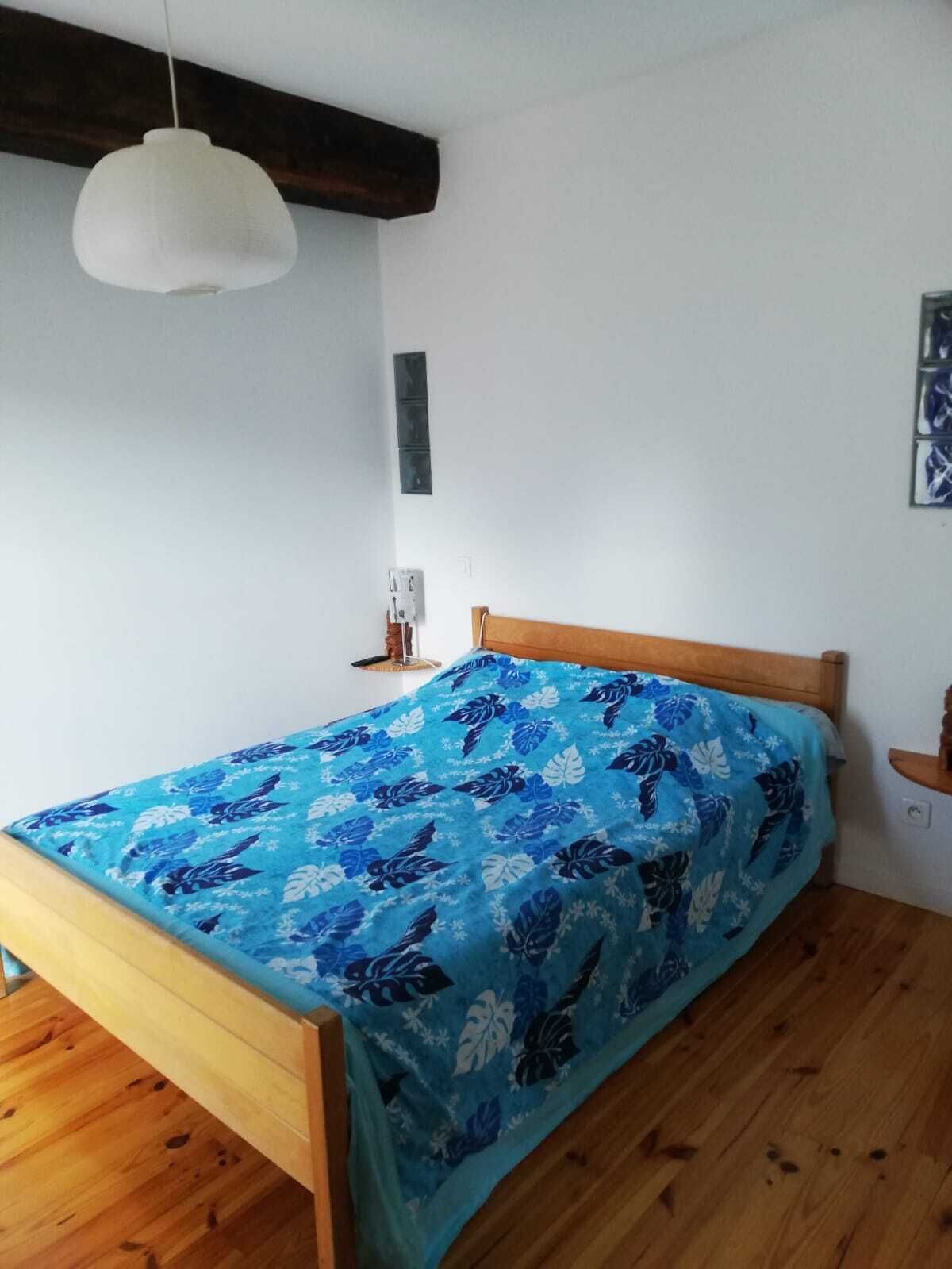 A bedroom with a wooden bed, a blue floral bedspread, a white pendant light, and a small bedside table.