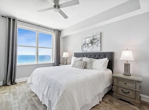 A bedroom with a large bed, two bedside tables with lamps, a ceiling fan, a window with a view, and a wall-mounted artwork.