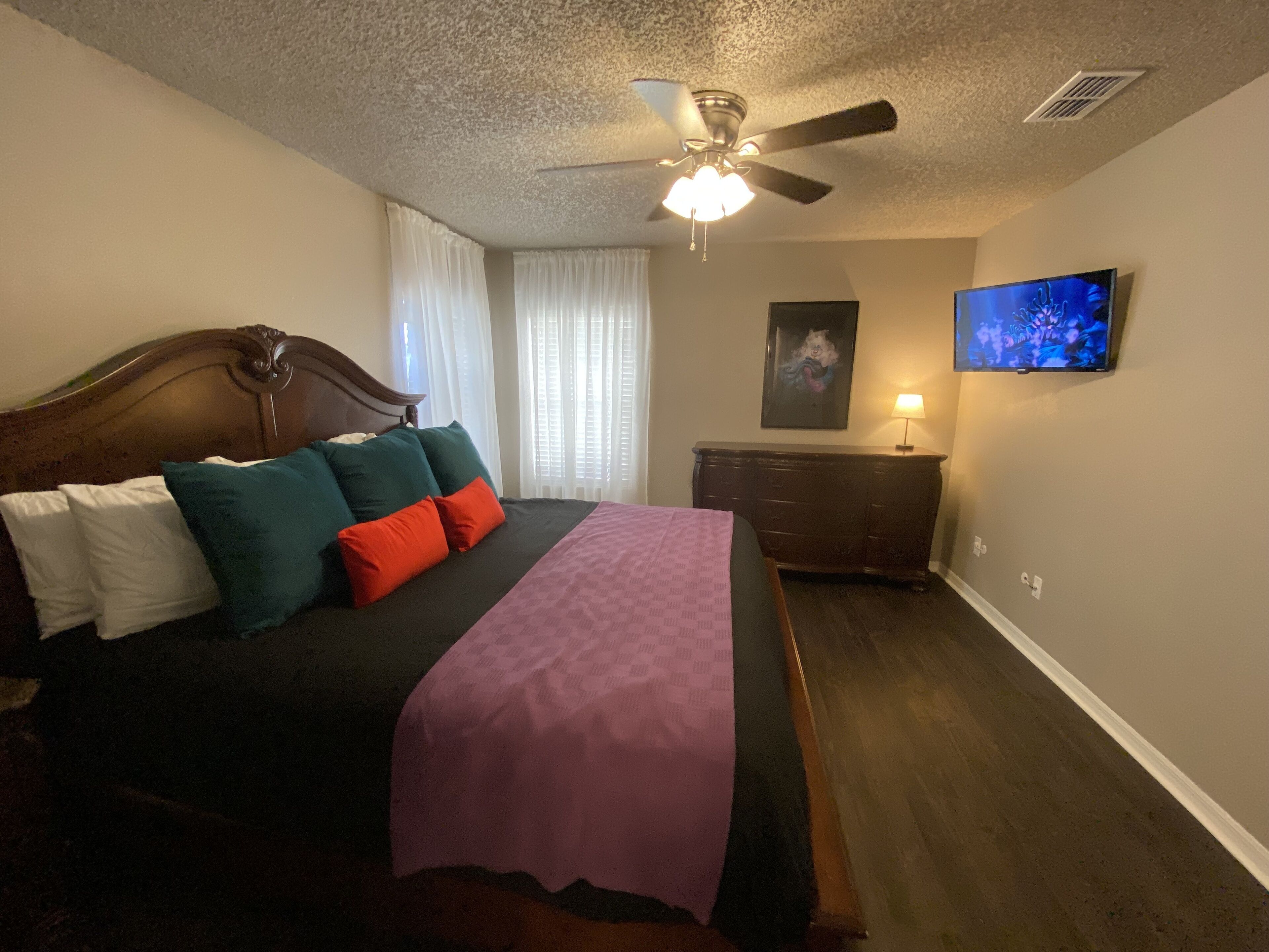 A bedroom with a bed, a ceiling fan, a dresser, a TV, and a window with curtains.