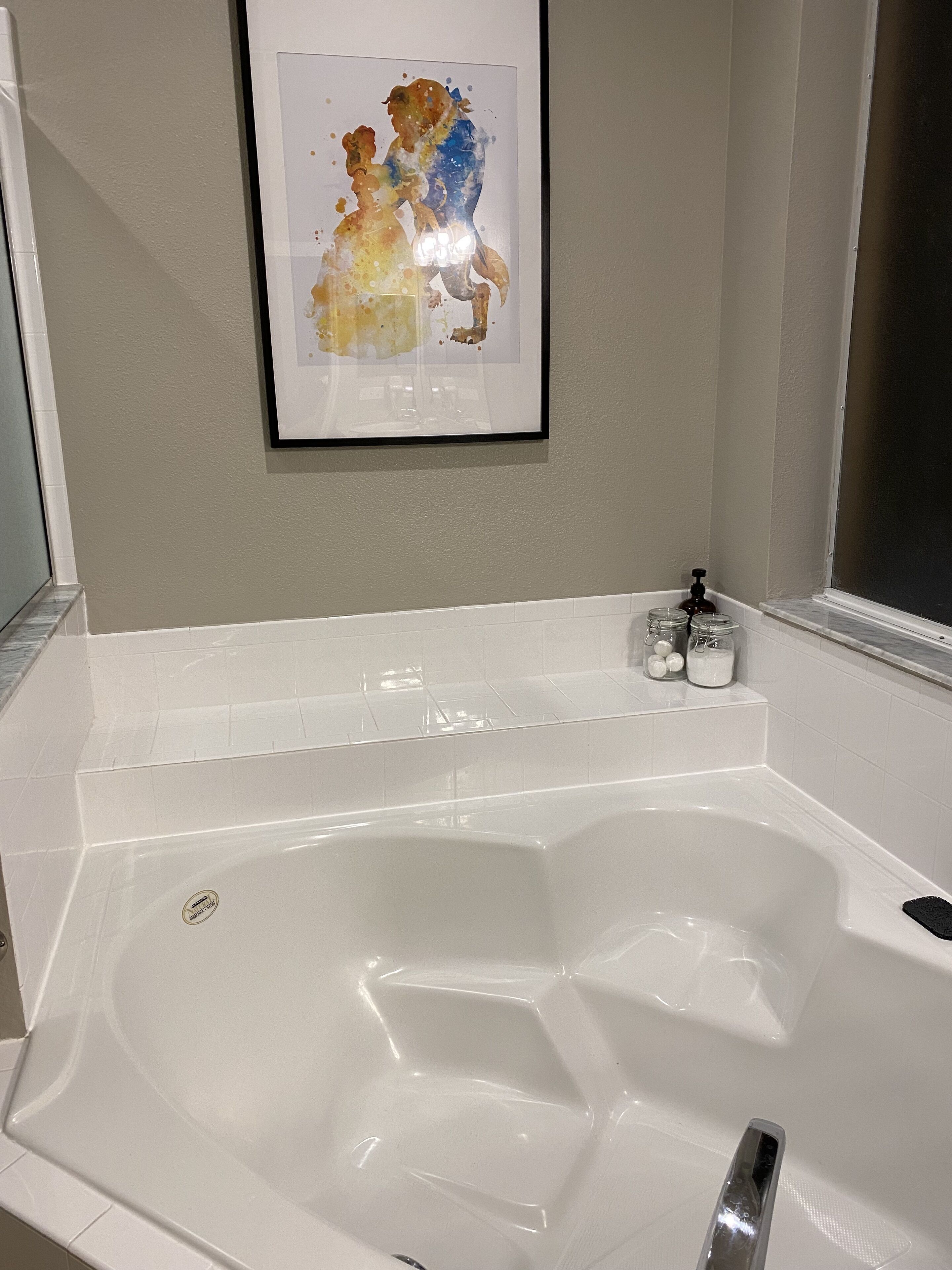 Ein Badezimmer mit einer großen weißen Badewanne, einem gerahmten Kunstwerk darüber und einem Fenster zur Rechten.