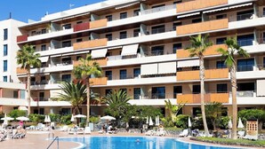 Ein mehrstöckiges Hotel mit Swimmingpool, Palmen und Liegestühlen.