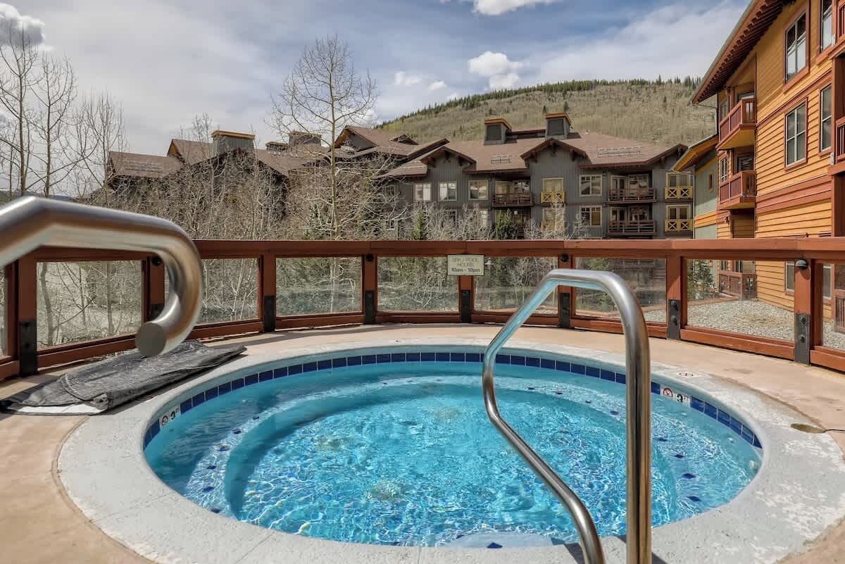 Un jacuzzi con una escalera metálica, rodeado por una terraza de madera y un paisaje de montañas.