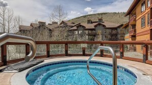 A hot tub with a metal ladder, surrounded by a wooden deck and a mountain backdrop.