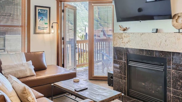 A modern living room with a leather sofa, a flat-screen TV mounted above a fireplace, and a large window with blinds.