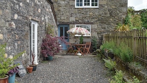 A stone cottage with a gravel driveway, a wooden fence, and a patio with a table and chairs.