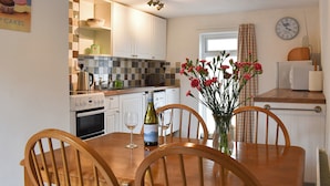 A kitchen with white cabinets, a wooden dining table with chairs, a clock on the wall, and a vase with flowers on the table.