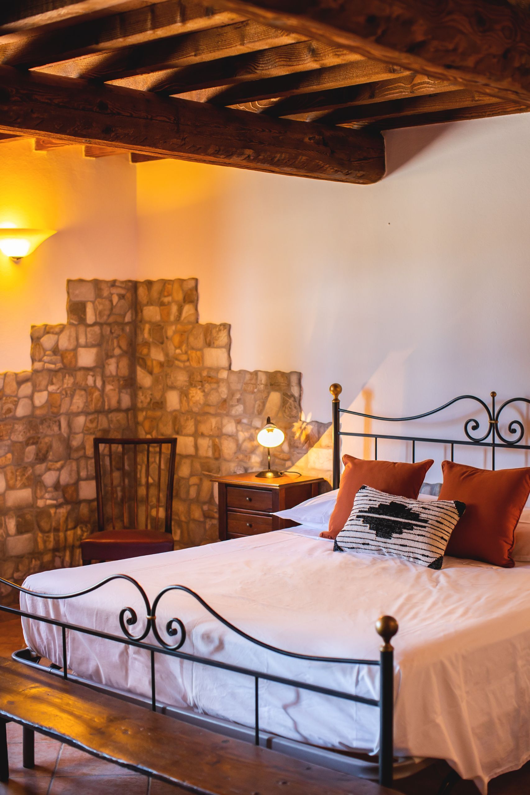 A bedroom with a stone wall, a wooden ceiling, a metal bed frame, a wooden bench, and a nightstand with a lamp.