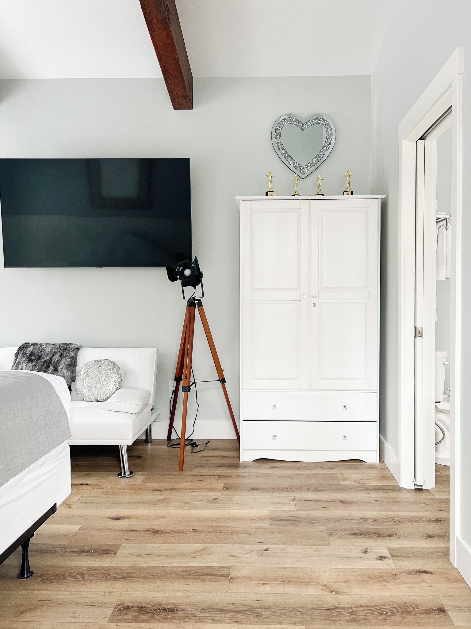A bedroom with a bed, a white wardrobe, a tripod, and a heart-shaped mirror on the wall.