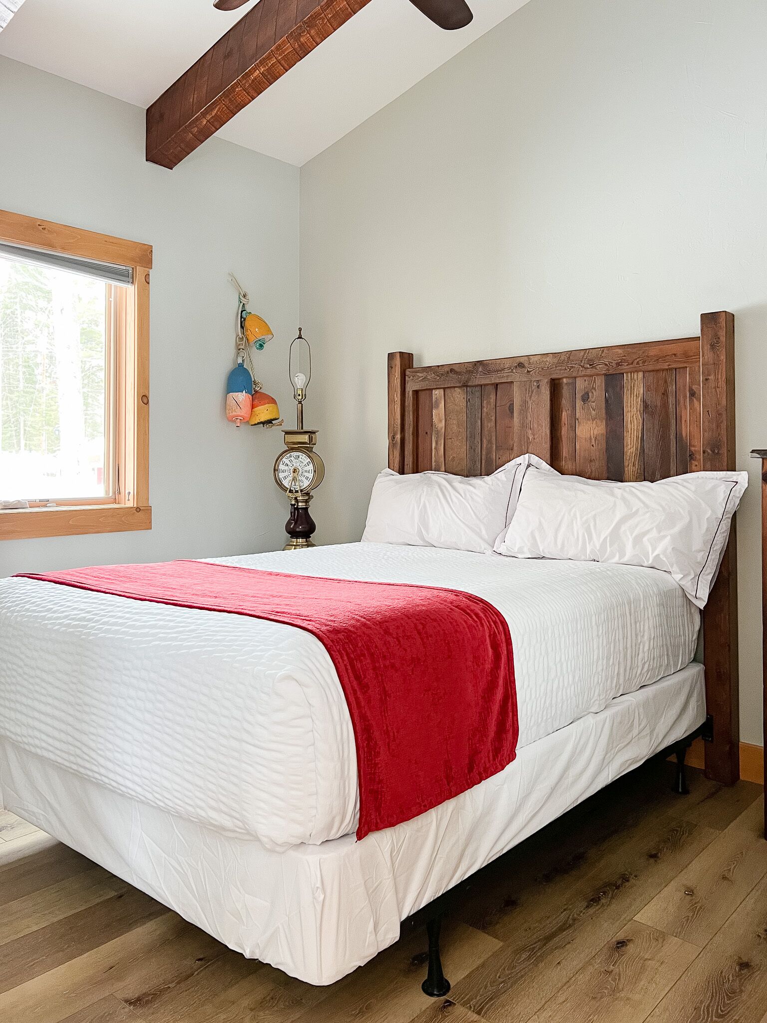 A bedroom with a wooden bed frame, white bedding, a red throw blanket, a nightstand with a lamp, and a window with a view of trees.