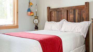 A bedroom with a wooden bed frame, white bedding, a red throw blanket, a nightstand with a lamp, and a window with a view of trees.