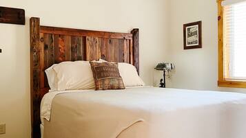 Un dormitorio con cabecera de madera, una cama grande con ropa blanca, piso de madera y una ventana con persianas.